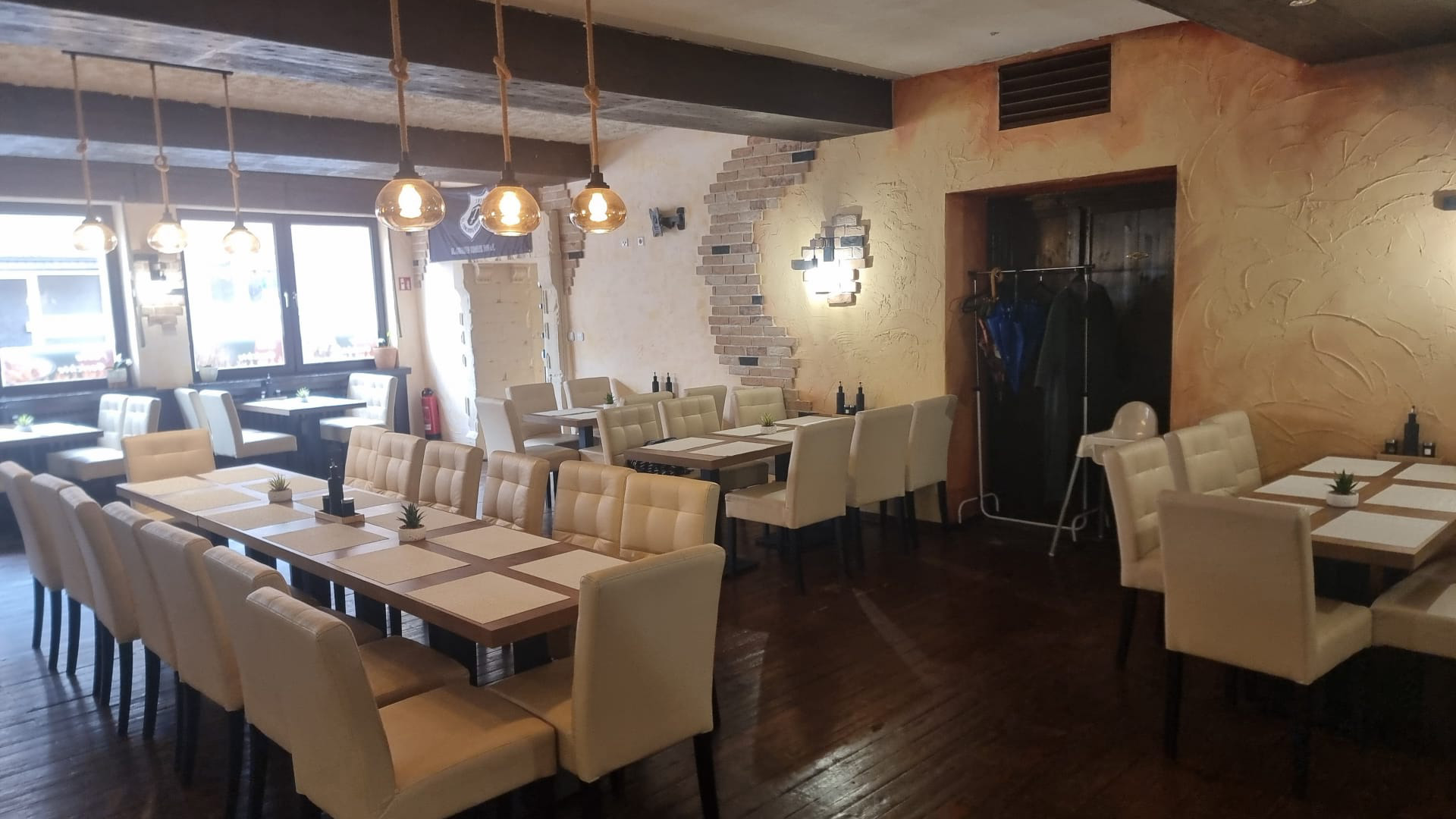 Leerer Speiseraum mit Holztischen, weißen Polsterstühlen und rustikaler Beleuchtung.